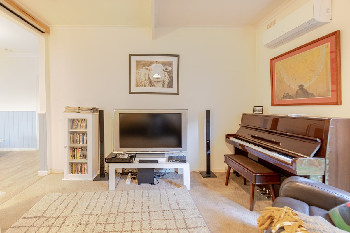 A spacious living area features a large television positioned on a white coffee table, with a black piano nearby. A warm light illuminates the room, which is adorned with tasteful art pieces and a soft area rug on the floor.