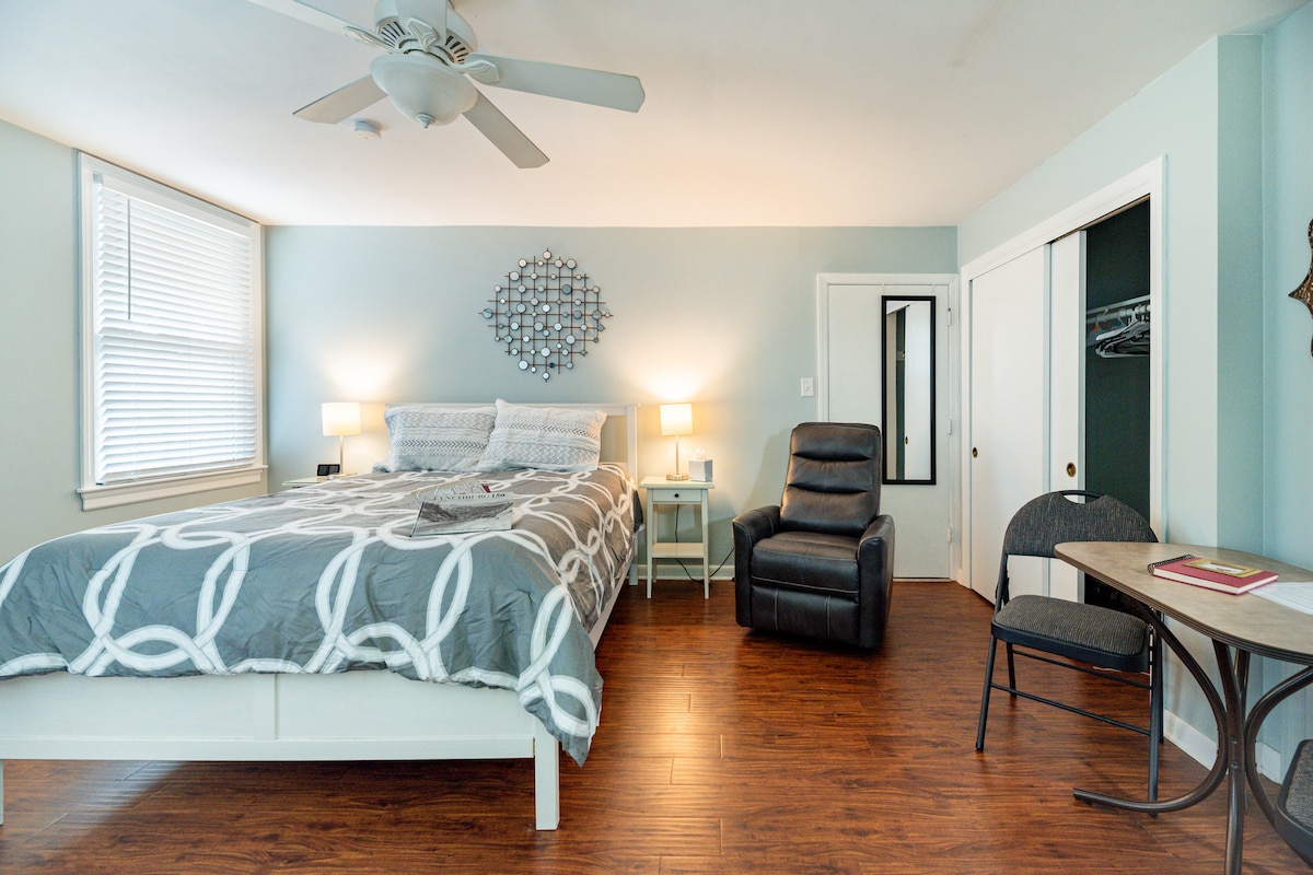 A spacious bedroom features a large bed with a patterned duvet and two bedside lamps. A comfortable recliner is placed nearby, accompanied by a small table and chair. Natural light enters through two windows, and a closet is accessible in the corner.