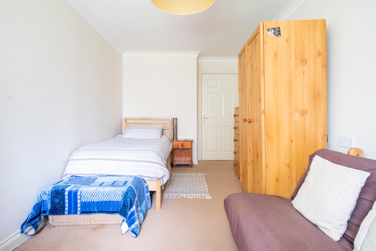 A spacious bedroom features a single bed dressed in light linens. A wooden wardrobe and chest of drawers provide ample storage. A cozy chair in a neutral tone is positioned nearby. Soft light is cast from a simple ceiling fixture, enhancing the room's openness.