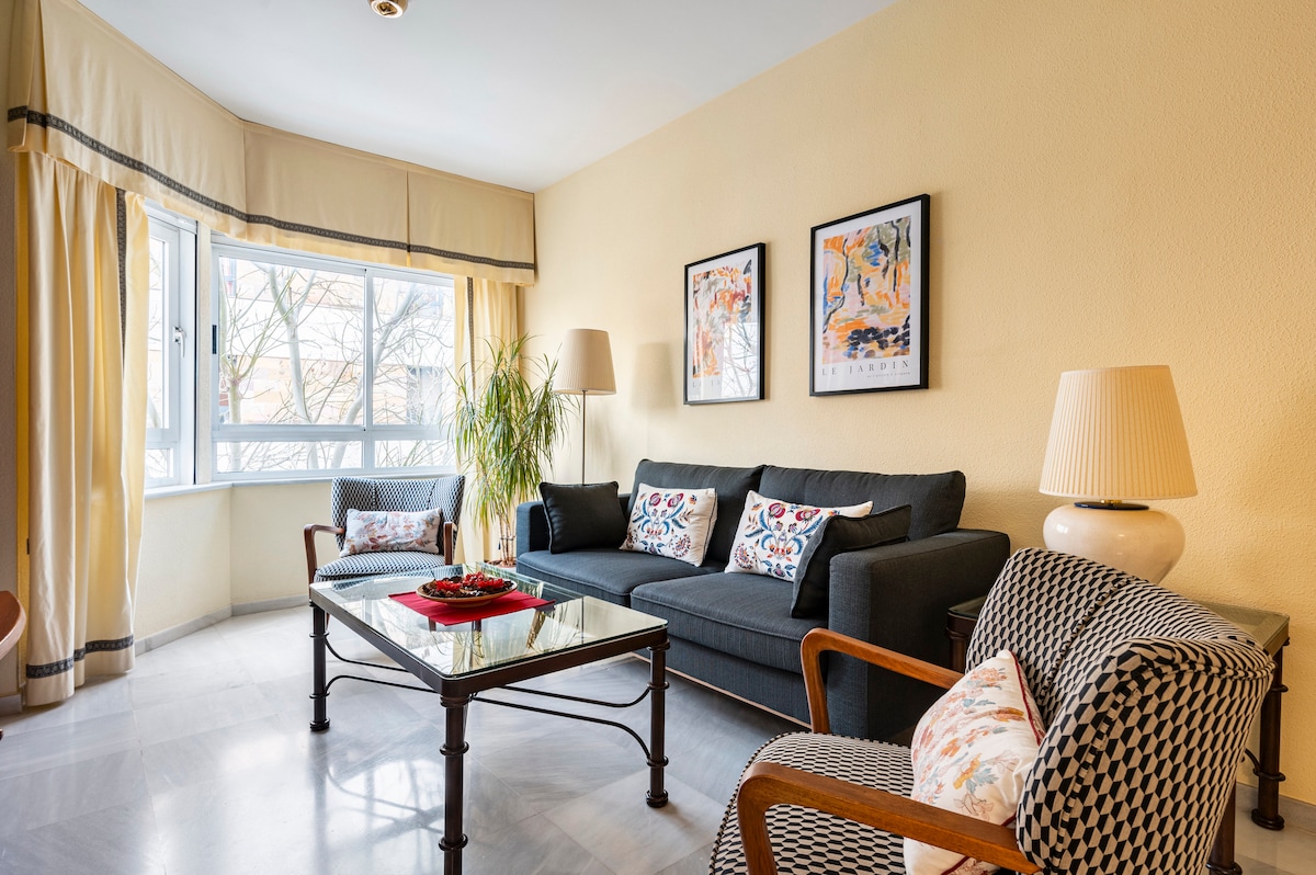 A bright living area features a comfortable dark sofa adorned with decorative pillows. Two framed art pieces are displayed on the wall, and a glass coffee table sits on patterned tile flooring. Natural light filters through large windows, surrounded by soft curtains.