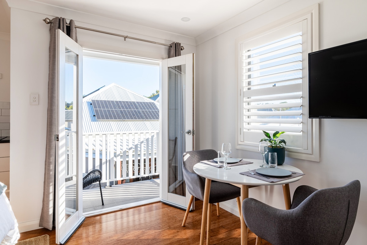 A bright dining area features a round table set for two, with modern gray chairs. Glass doors open to a balcony, allowing natural light and fresh air. The view includes rooftops with solar panels and a glimpse of the outdoors, enhancing the spacious feel.