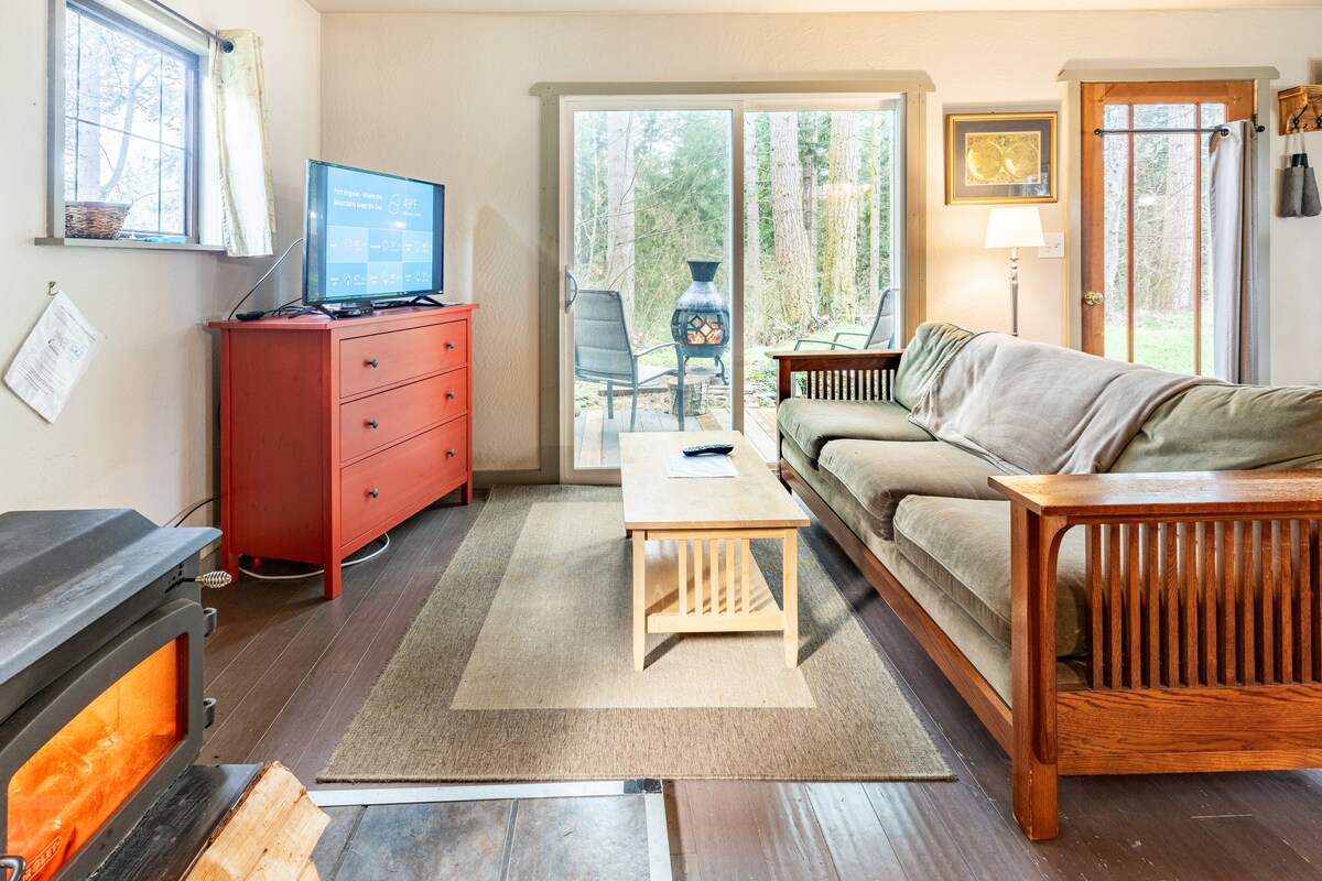 A comfortable living area features a large sectional sofa with neutral-toned fabric, a wooden coffee table, and a red dresser with a television. Natural light fills the space through a large sliding door, which opens to an outdoor sitting area with a fire feature.
