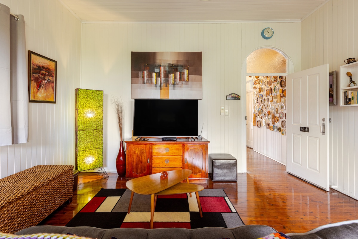 A spacious living area features a large flat-screen TV and a wooden sideboard, complemented by a decorative floor lamp. Colorful rugs cover the polished wooden floor, and light enters through large windows with sheer curtains, enhancing the room's airy feel.
