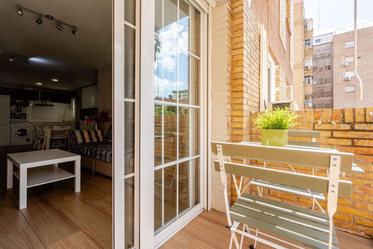 A sunlit balcony features a small table and two chairs, surrounded by warm brick walls. The adjoining open door leads to a cozy living area with modern furnishings. Gentle sunlight filters in, creating a bright and airy ambiance throughout the space.