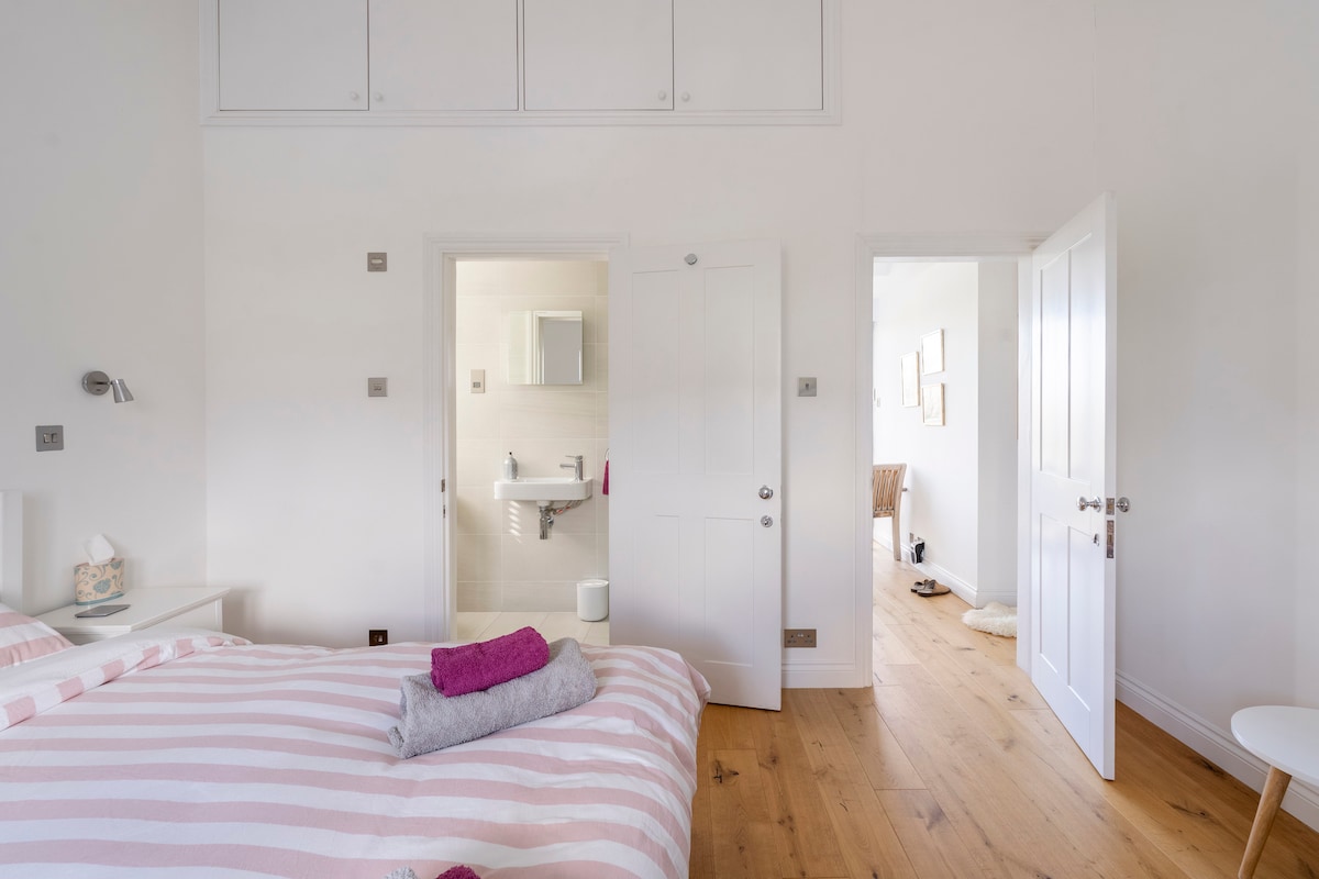 A bright double bedroom features striped bedding and neatly arranged towels. Doors lead to an ensuite bathroom and a hallway. Light-colored walls and wooden flooring enhance the spacious feel, while minimalistic decor adds to the serene atmosphere.