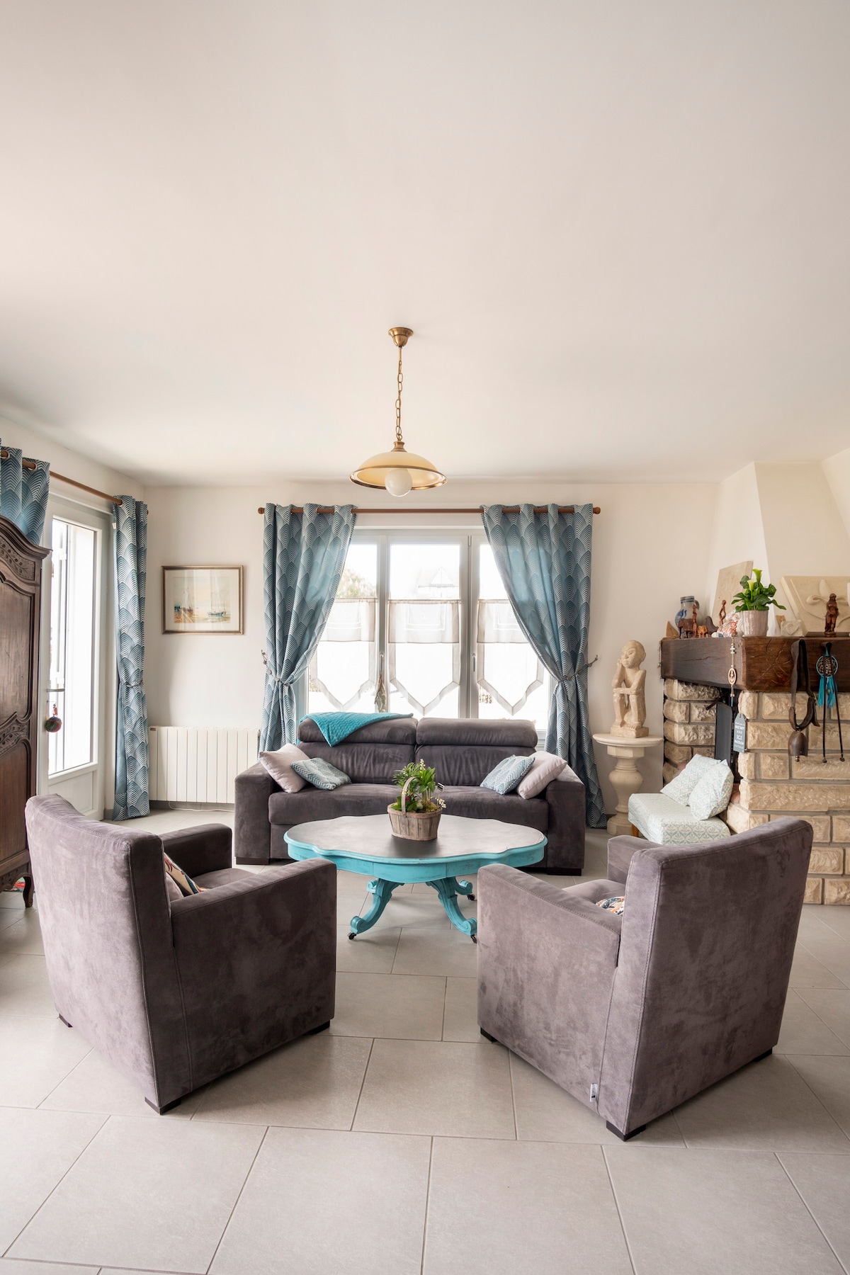 A cozy living area features a round table surrounded by four upholstered chairs. Large windows provide natural light, framed by sheer blue curtains. A decorative fireplace and various plants enhance the space, while neutral-toned walls contribute to a calm ambiance.