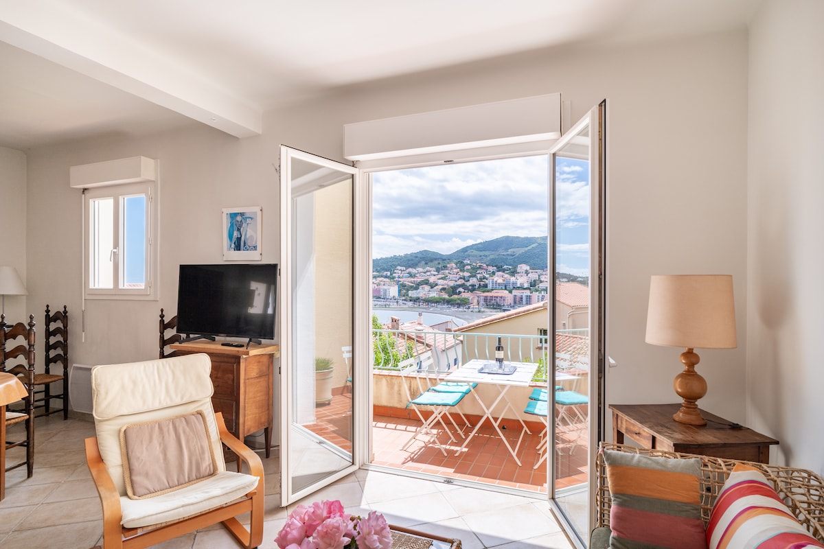 A bright living area features large glass doors that open to a terrace, showcasing views of the harbor and surrounding hills. A light-colored chair and wooden furniture are present, along with a small table and two chairs on the terrace. Natural light fills the space.