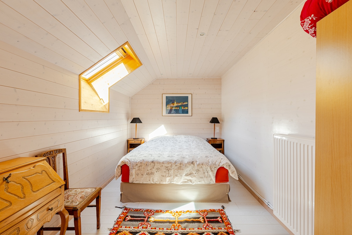 A bright bedroom features a spacious bed with light bedding, flanked by two bedside tables and lamps. A decorative rug lies on the floor, and a small chair sits next to a wooden cabinet. Natural light enters through a skylight and side window.