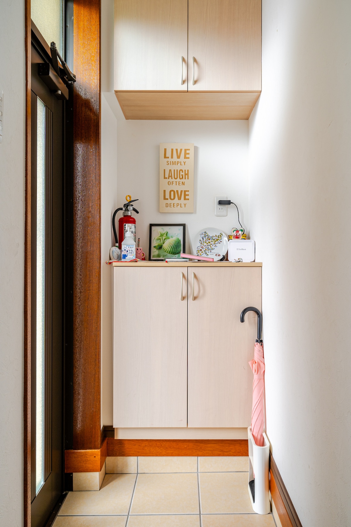A compact entryway features light wood cabinetry providing storage above and below. A fire extinguisher and decorative items are arranged on the counter, while an umbrella stand holds a pink umbrella. Natural light filters through the glass front door, enhancing the welcoming atmosphere.