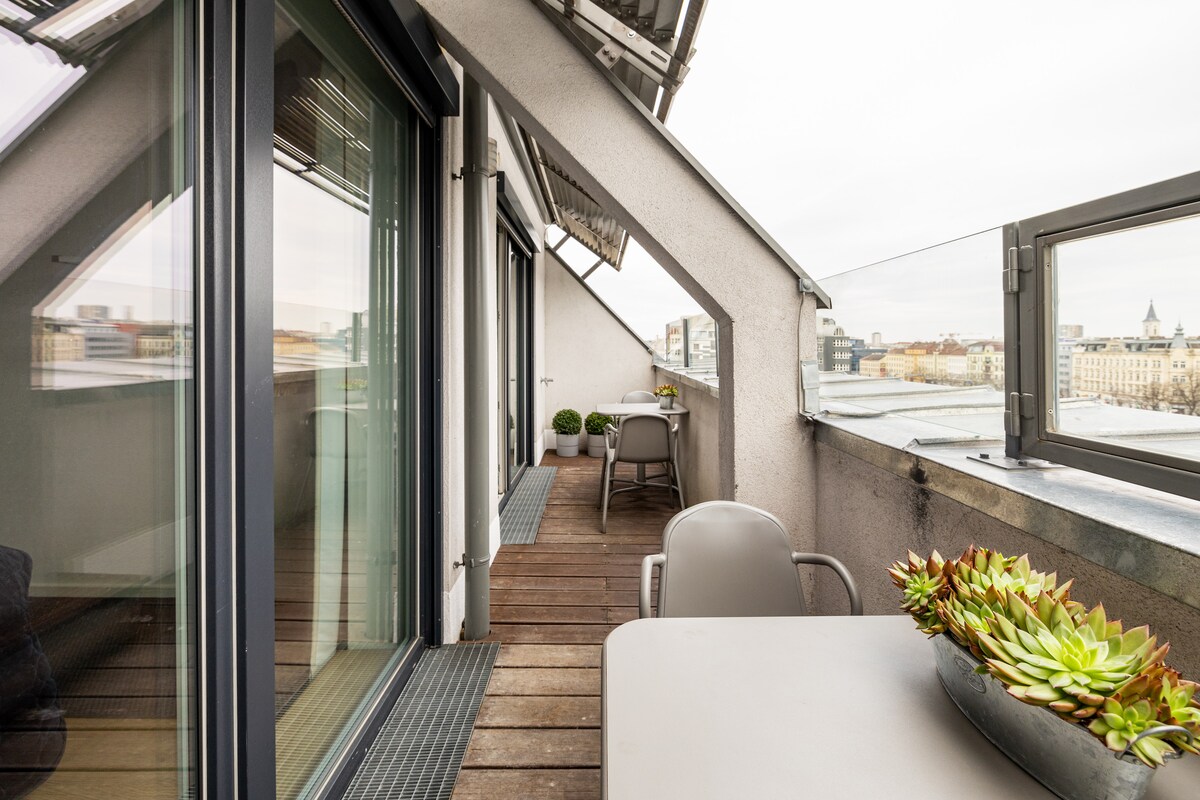A north-facing terrace is displayed, featuring wooden flooring and minimalist outdoor furniture. Potted plants line the edges, while a table is set for casual dining. Expansive glass doors provide panoramic views of the surroundings, enhancing the connection between indoor and outdoor spaces.
