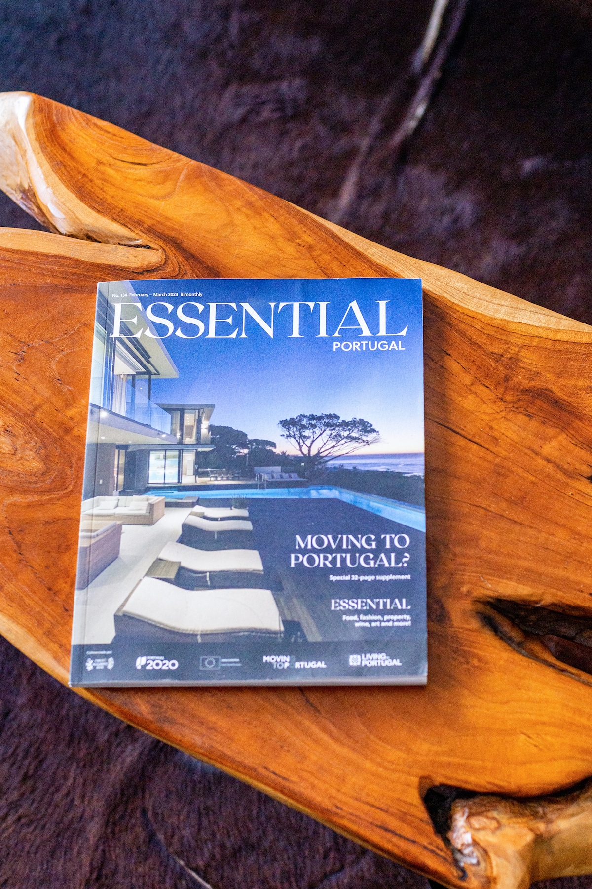 A magazine titled 'Essential Portugal' is placed on a textured wooden table. The magazine features a sleek cover image of modern architecture beside a serene landscape, suggesting themes of relocation and lifestyle in Portugal.