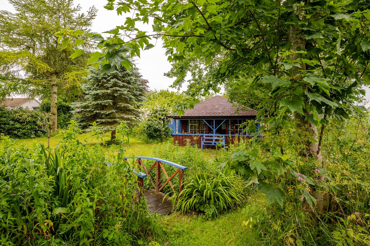 A wooden summerhouse is nestled among lush greenery, surrounded by tall trees and blooming plants. A small red bridge crosses over a walkway leading to the entrance, inviting guests to explore the serene garden setting.