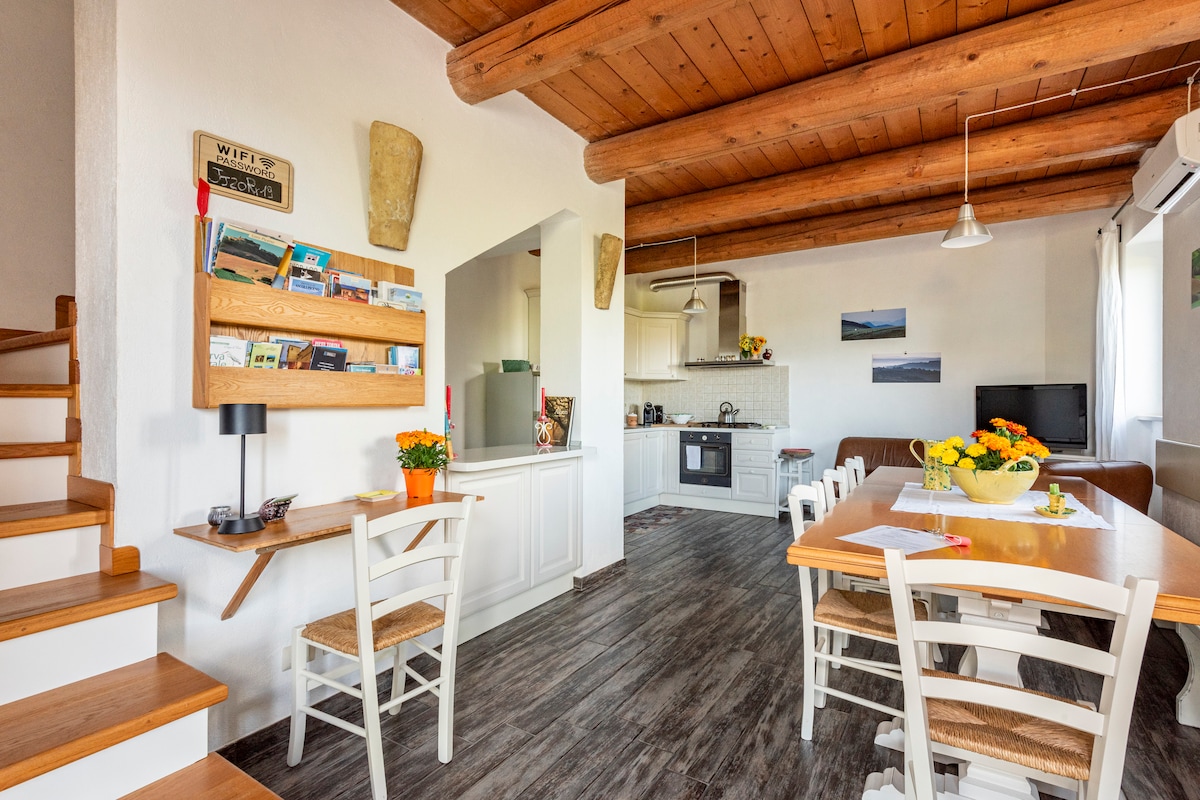 A bright and airy living area is depicted, featuring wooden beams and a spacious kitchen equipped with modern appliances. A dining table with chairs sits prominently, surrounded by decorative flowers. A small staircase leads to the upper level, enhancing the cozy atmosphere.