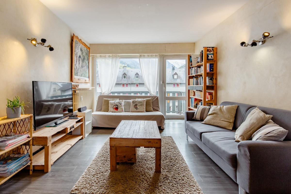 A spacious living area showcases a cozy sofa and a light-colored couch opposite a wooden coffee table. A large window allows natural light to fill the room, with soft curtains framing the view. A bookshelf filled with various books adds character to the space.