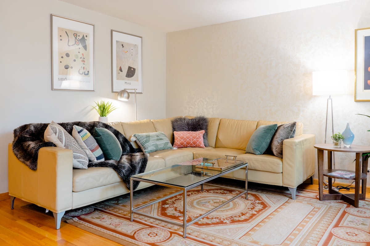 A spacious living area showcases a comfortable L-shaped leather sofa adorned with decorative pillows. A glass coffee table is positioned on an elegant area rug, while framed art pieces decorate the walls. A floor lamp provides soft lighting next to a small side table with a decorative plant.