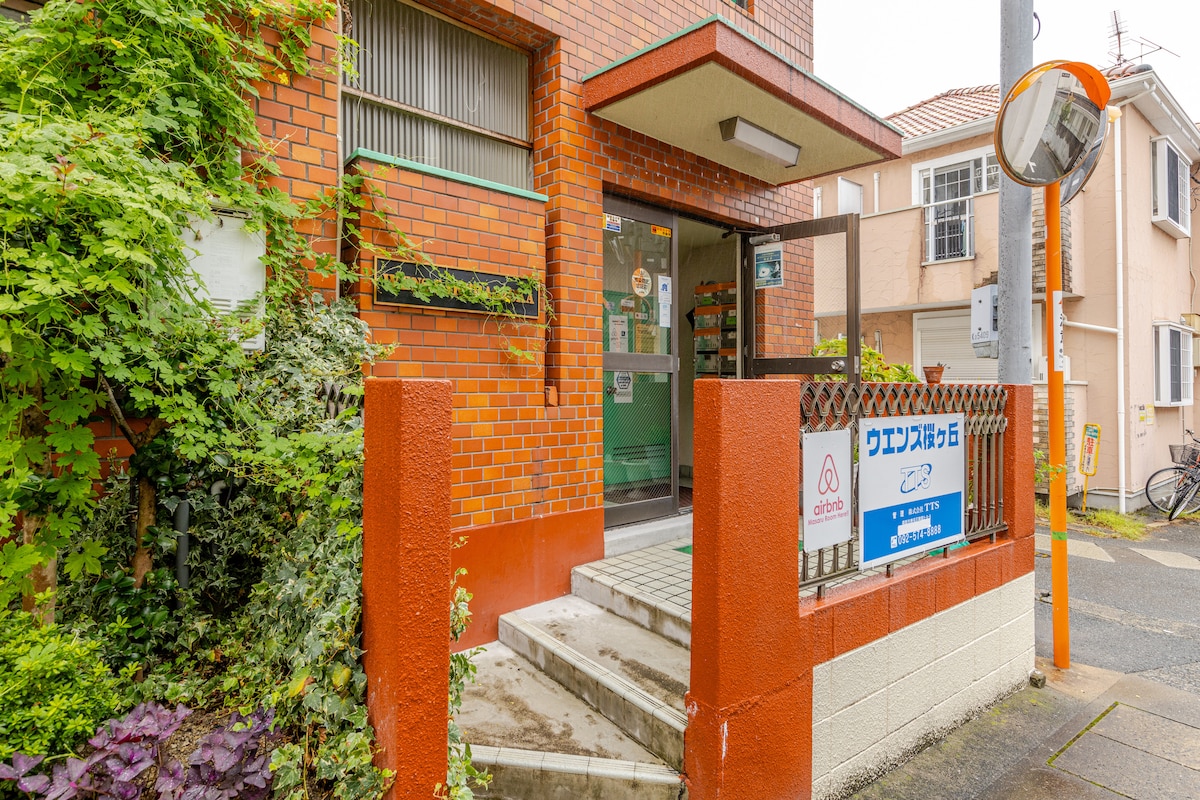 The entrance to the apartment building features a brick facade and a set of stairs leading up to the entryway. Green vegetation and signage mark the doorway. A bicycle is visible nearby, with a street sign indicating the area.