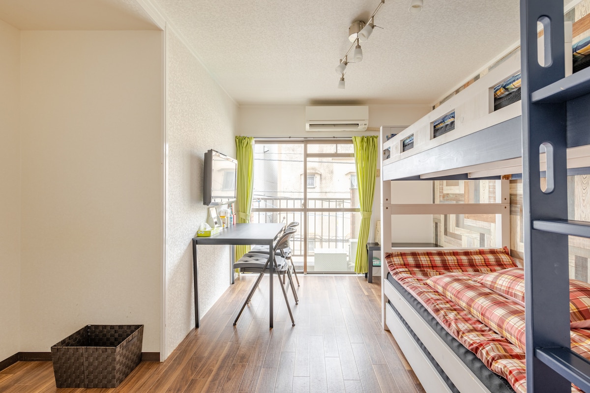 A bright, open room features a lofted bunk bed with a red plaid covering. A desk with two chairs is positioned near a large window, adorned with green curtains. A dark basket is located on the wooden floor, contributing to the room's functional layout.