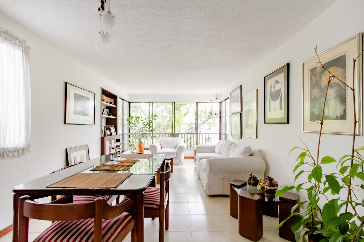 A bright living space features a dining table with patterned chairs, surrounded by framed artwork on the walls. Soft beige sofas are positioned near large windows, allowing natural light to fill the room. Green plants add a touch of nature to the area.