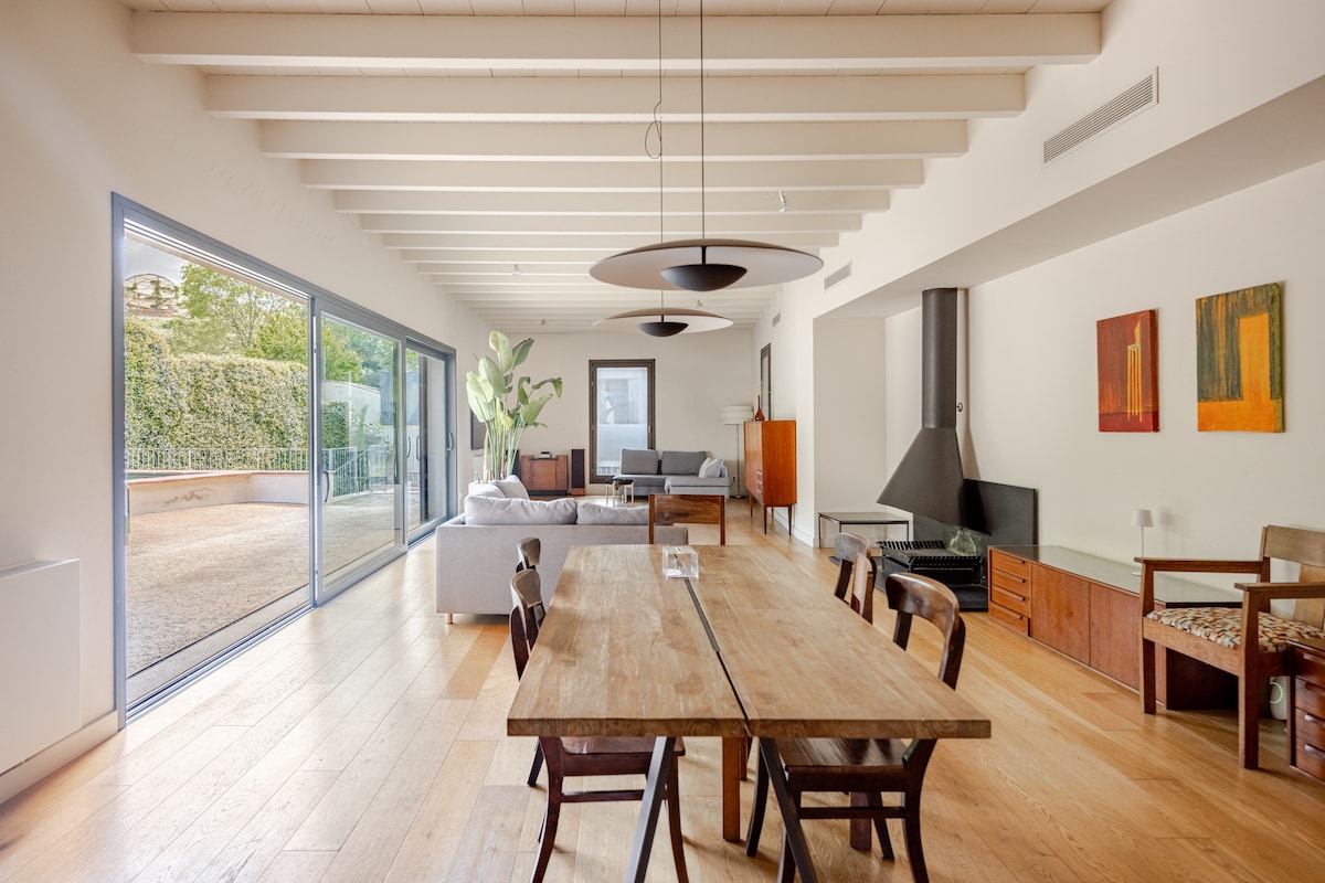 A spacious living and dining area features wooden flooring and high ceilings with exposed beams. Large glass doors offer views of the patio while allowing natural light to fill the room. A wooden dining table is centrally located, surrounded by wooden chairs, and a cozy seating area is visible to one side.