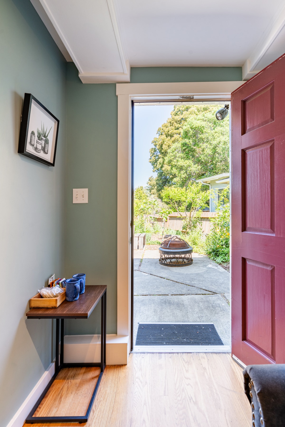 The entryway features a red door that opens to a bright outdoor area. A small table with items is positioned against a wall, while a view of a landscaped garden and outdoor seating is visible through the doorway.