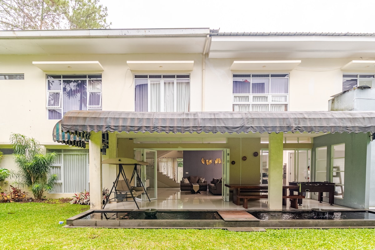 The exterior view showcases the spacious villa with large windows and a covered outdoor area. A tranquil water feature reflects the surrounding greenery, while a shaded seating area provides a comfortable spot to relax. The well-maintained lawn complements the serene setting of the villa.