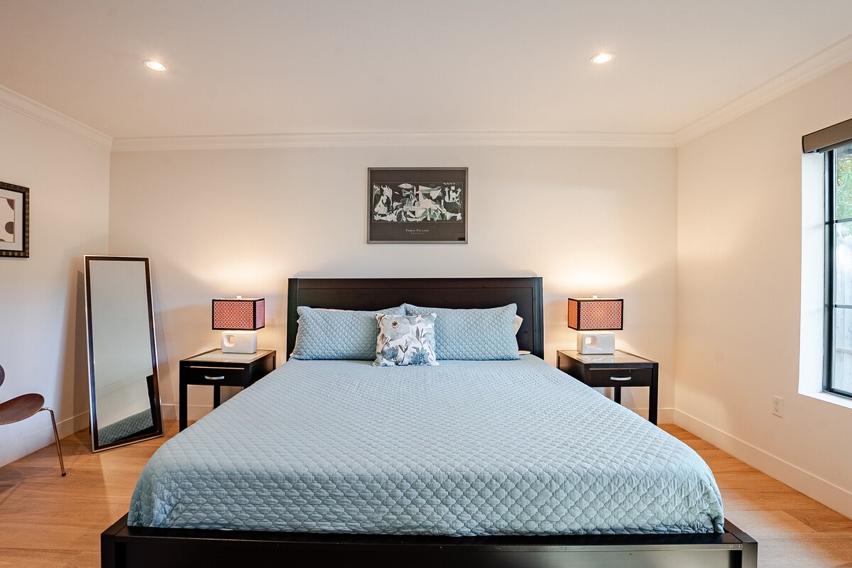 في الصورة: سرير بحجم كبير، ومنضدتان ليليتان مع مصابيح في كل منهما، مرآة كاملة الطول