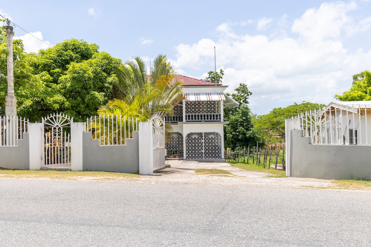 The exterior of a two-story cottage is seen, featuring intricate wrought iron gates and a spacious, inviting front porch. Lush green trees frame the property, with a hint of the surrounding countryside visible in the background under a bright sky.