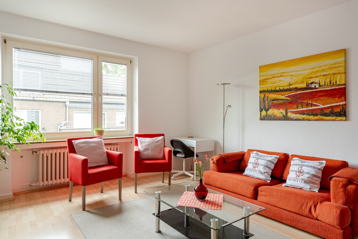 A bright living area features a glass coffee table surrounded by an orange couch and two coordinating armchairs. Natural light fills the space through a large window, while a piece of art depicting a countryside landscape adorns the wall.