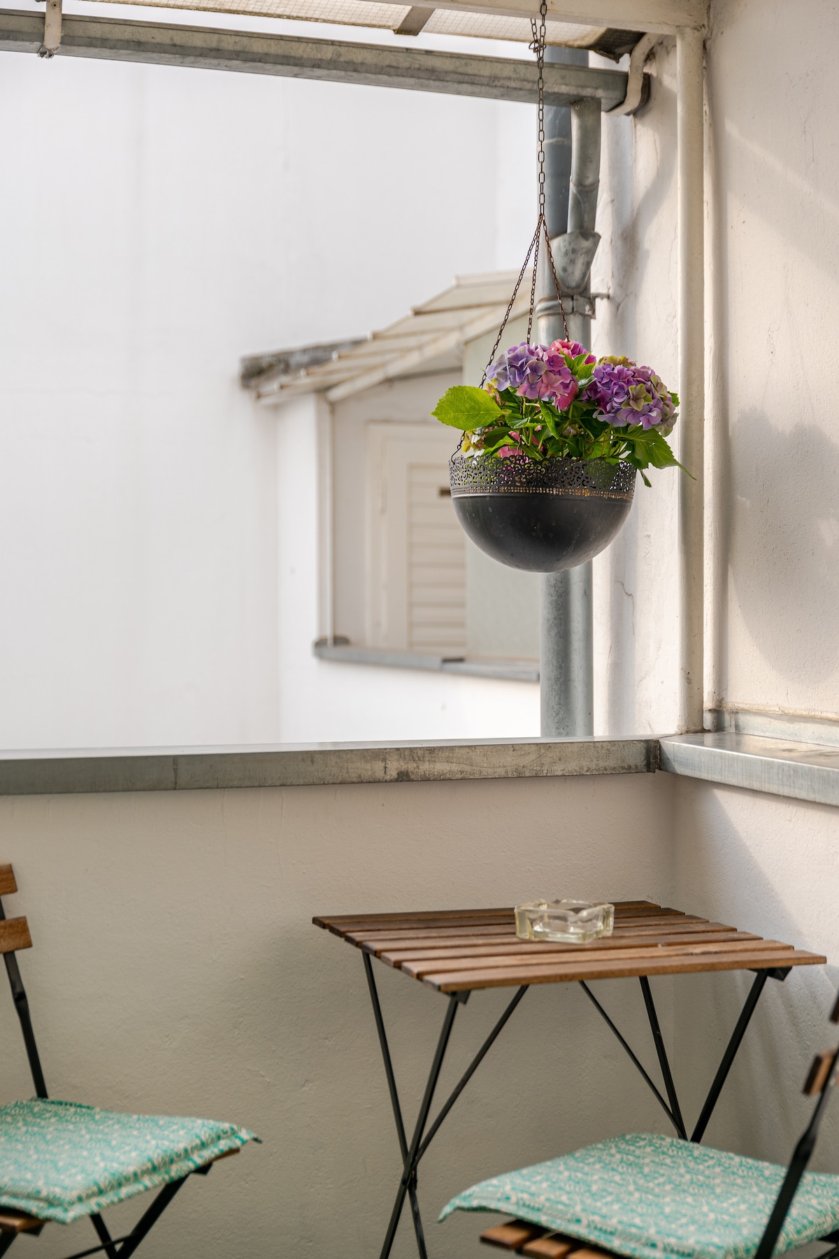A small balcony features a wooden table paired with two folding chairs. A hanging planter with vibrant purple flowers adds a touch of color, while a glass ashtray sits atop the table. The backdrop consists of a light-colored building.