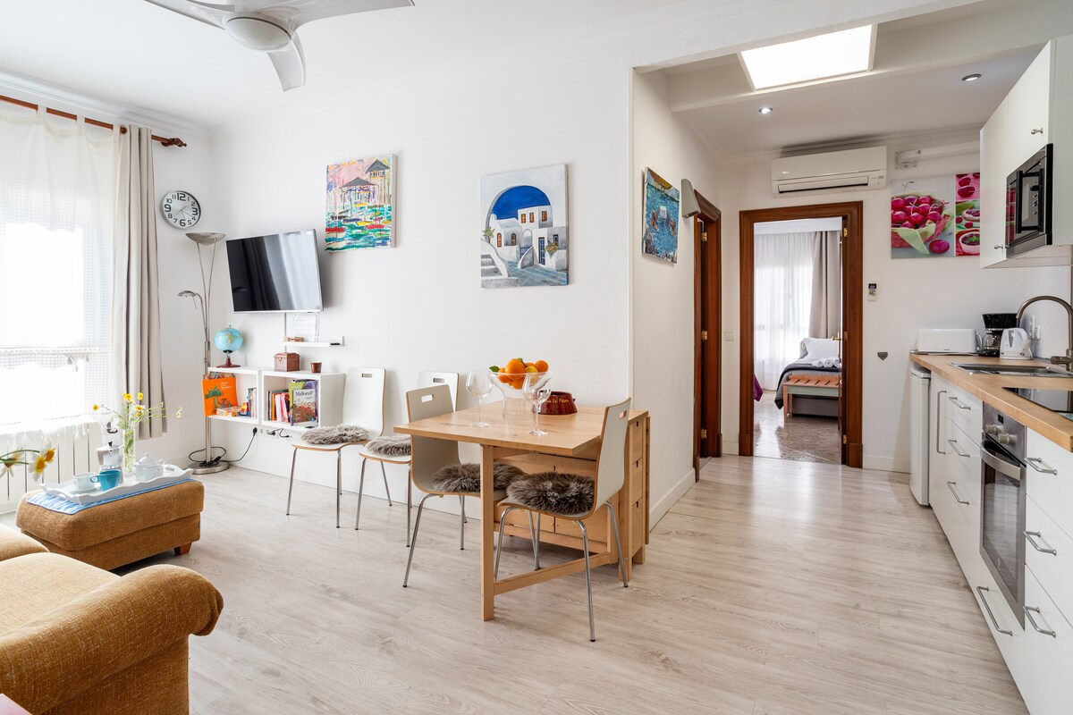 An open-plan living area features a dining table set for four, with a modern kitchen in the background. Natural light floods the space through large windows adorned with sheer curtains. A comfortable seating area offers a cozy place to relax, complemented by colorful wall art.