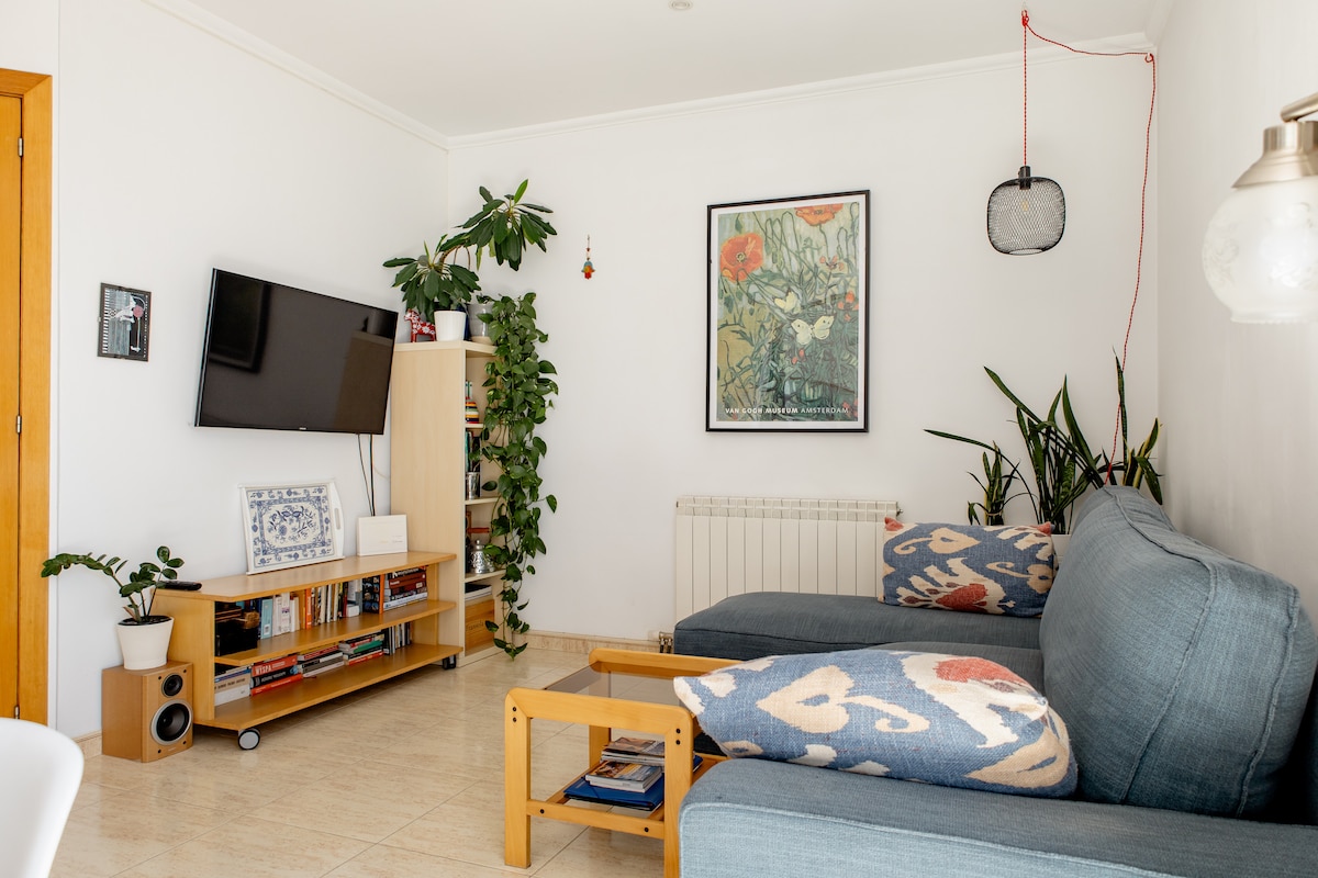 A bright living area features two comfortable blue sofas, accented with patterned cushions. A wall-mounted television is positioned above a media console, which holds books and decorative items. Green plants enhance the space, while light-colored stone flooring contributes to a welcoming environment.