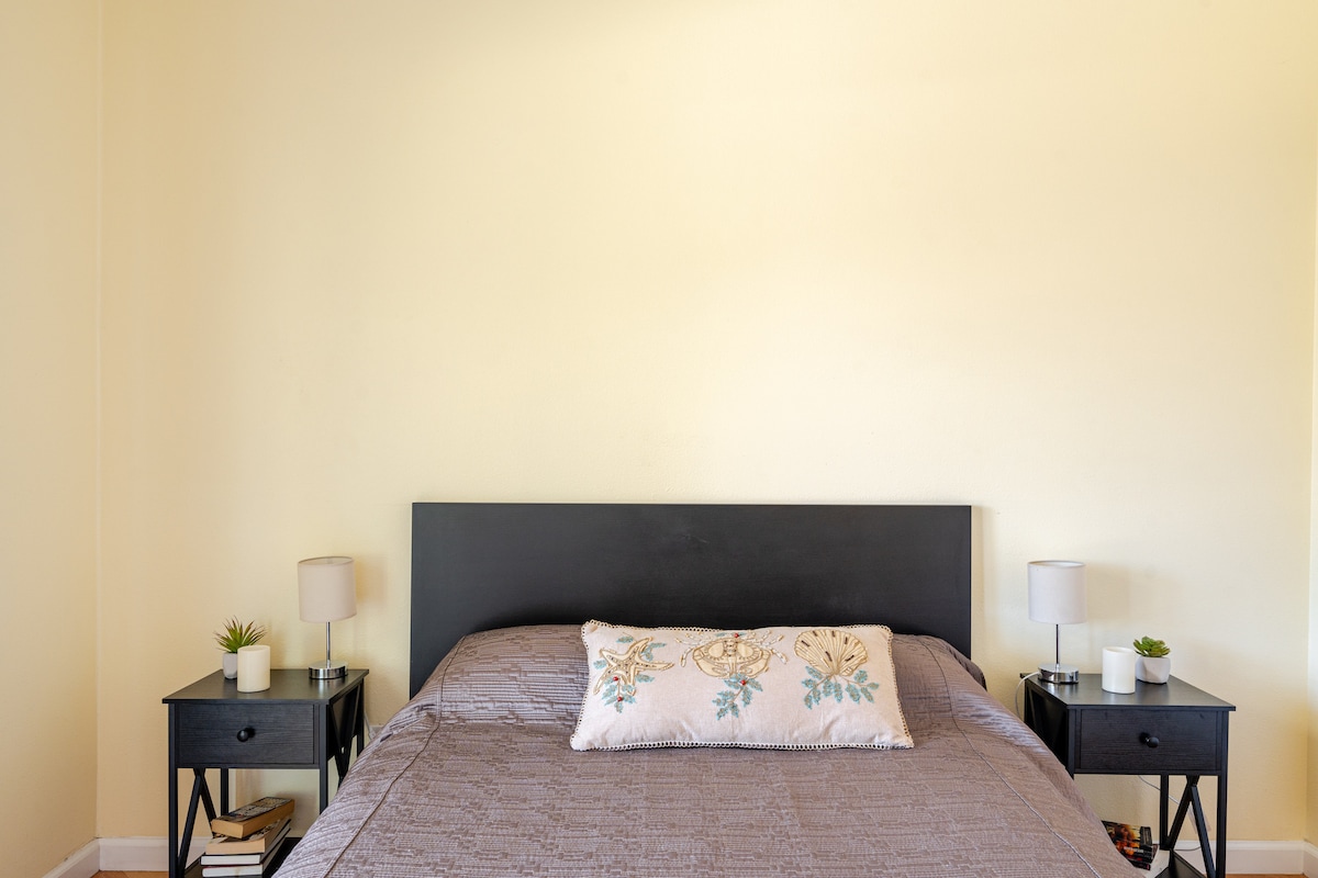 A cozy sleeping area features a spacious bed with an ornamental throw pillow resting against a simple black headboard. Flanking the bed are two nightstands, each equipped with a lamp and a small plant. Light yellow walls provide a calming backdrop.