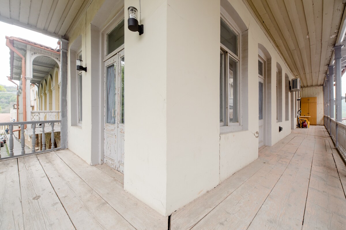 The spacious balcony showcases a wooden floor with a light finish, surrounded by walls painted in soft tones. Natural light enters through several large windows, creating an airy ambiance. The view offers glimpses of the surrounding architecture and the outdoors.