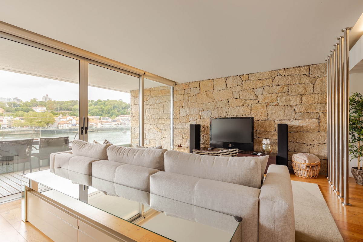 A spacious living area features a light-colored sectional sofa facing a flat-screen television. A glass coffee table rests in front, while large windows showcase views of the waterfront. Natural light fills the room, and a stone accent wall adds texture to the modern design.