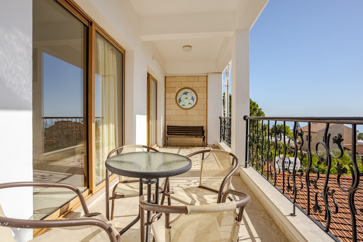 A balcony area is captured, featuring a glass-topped round table surrounded by four metal-framed chairs. A wooden bench is positioned against the wall, with a scenic view of the surrounding landscape visible through the sliding glass doors.