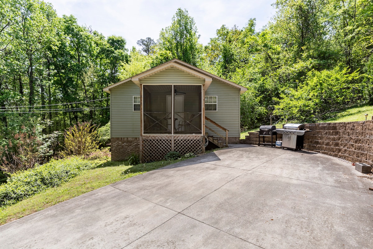 A serene exterior view of a house surrounded by lush greenery. A large screened porch is featured at the front entry, with a concrete driveway leading up to the entrance. Outdoor grilling equipment is positioned nearby, enhancing the outdoor living space.