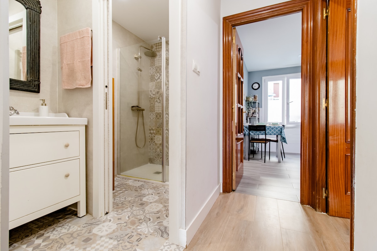 A view from the hallway showcases a shared bathroom area featuring a modern shower. A white vanity with a mirror is positioned beside the shower. The door to a dining area with a blue table and chairs is visible at the end of the hall, leading to natural light.