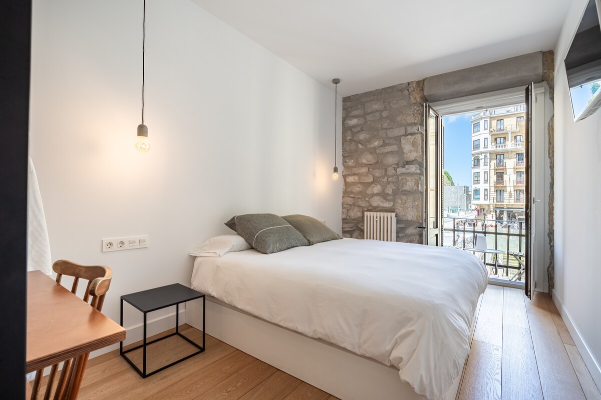 A bright bedroom features a spacious bed with neutral bedding, flanked by two soft pillows. A small desk and chair are visible next to the bed. Natural light enters through a balcony door, revealing views of the street. A stone wall adds a touch of character.