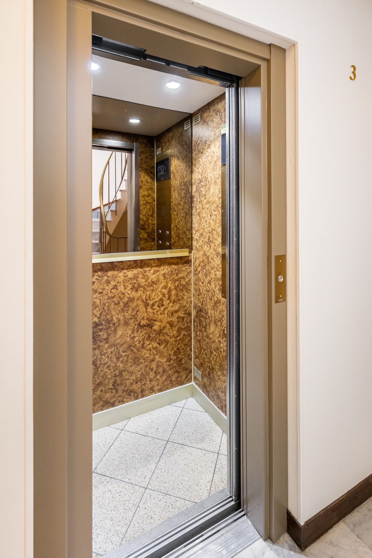 An elevator with a sleek design features a mirrored interior, reflecting warm wood paneling and soft overhead lighting. The ground is made of neutral-colored tiles, and a spiral staircase is visible in the background, providing easy access to upper levels.
