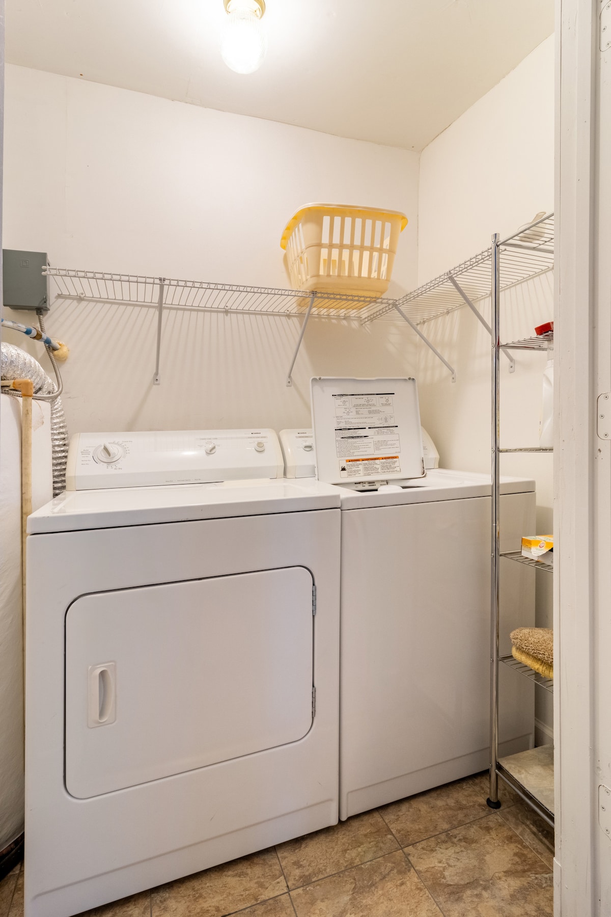 A compact laundry area is equipped with a white washing machine and dryer, accompanied by a drying rack and a wire shelving unit. A yellow laundry basket sits atop the shelf, and a tiled floor adds functionality to the space.