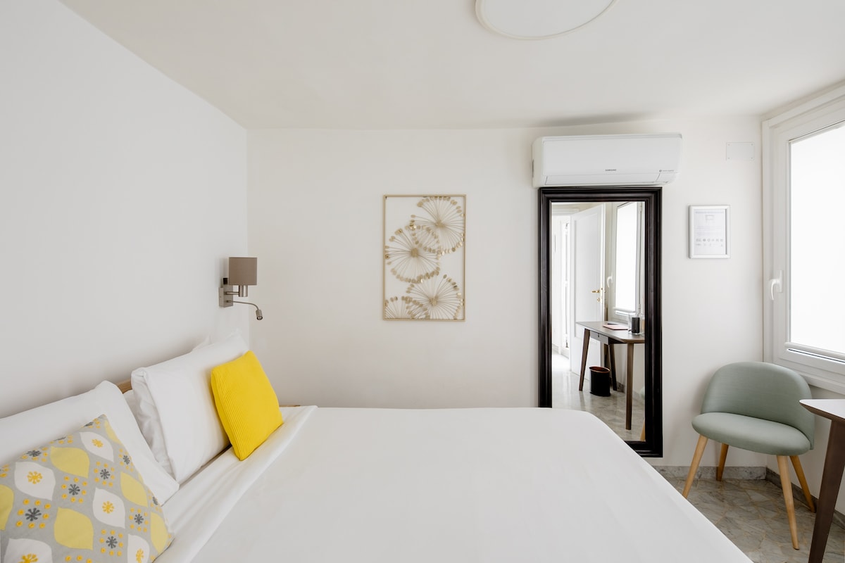 The bedroom features a spacious bed adorned with a yellow accent pillow and a decorative cushion. A mirror reflects natural light, and a contemporary chair is positioned beside a small table. Light walls and soft decor create a calming space.