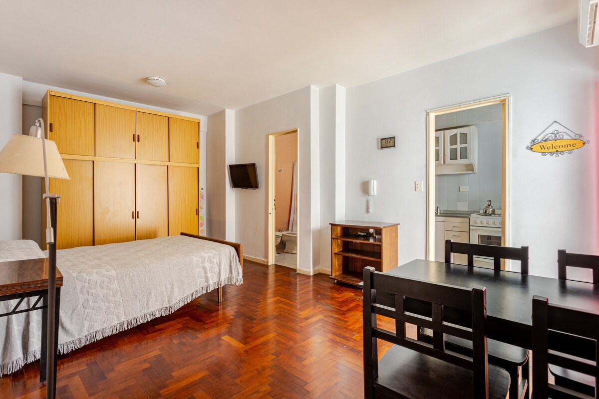 A studio apartment features a spacious bed and a wooden dining table accompanied by four chairs. A large wardrobe stands against the wall, while a smart TV is mounted nearby. The kitchenette is visible through an open door, providing essential amenities.