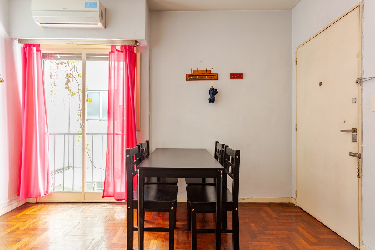 A dining area features a black rectangular table surrounded by six dark chairs. Large windows, adorned with sheer pink curtains, allow natural light to fill the space. A wall-mounted coat rack is visible, along with a simple door leading outside.