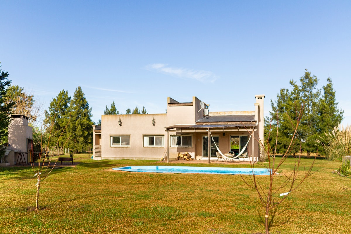 A private outdoor space features a house with large windows, surrounded by green grass and trees. A swimming pool is visible in the foreground, inviting relaxation, while a shaded area with seating offers a tranquil spot for gatherings.