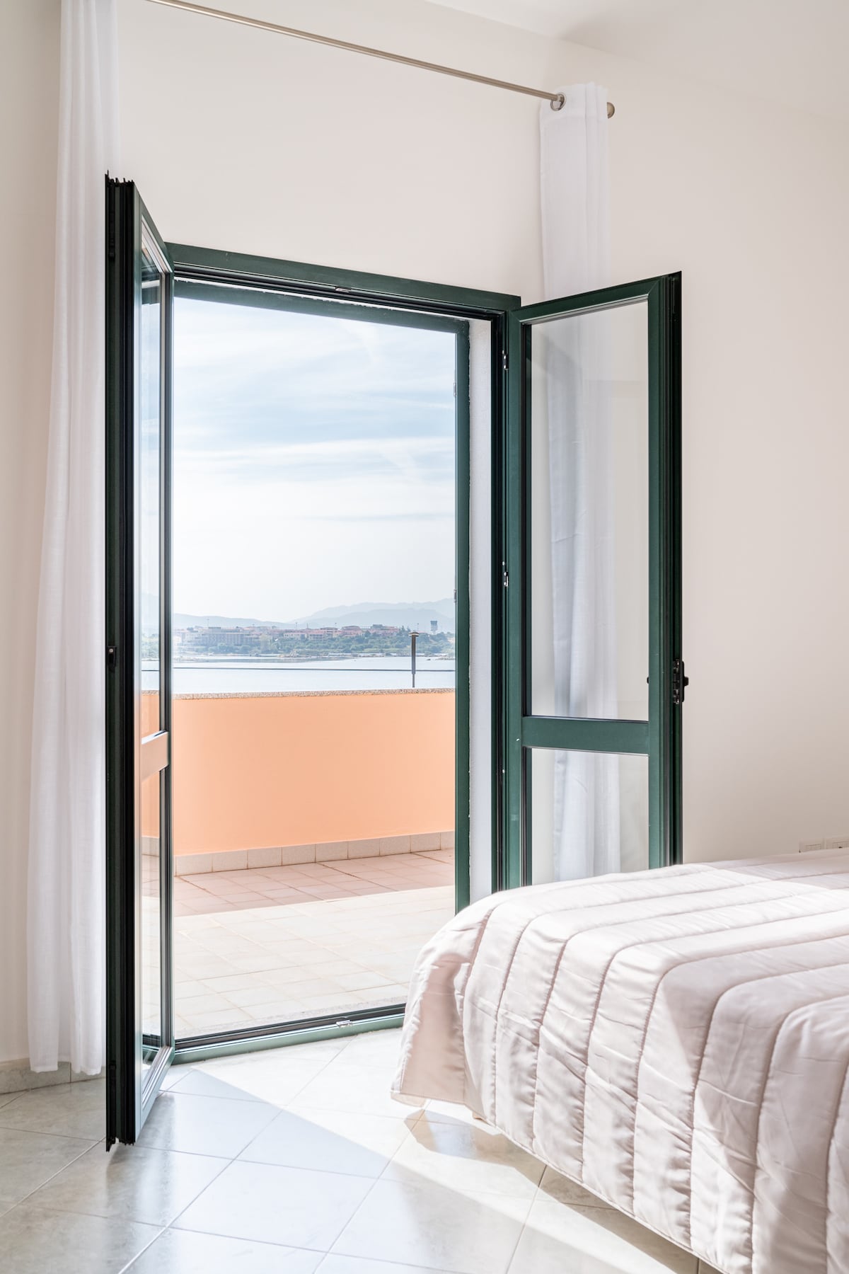 A bright bedroom features a spacious bed with light bedding. Large double doors open to a balcony, providing unobstructed views of the surroundings. Soft natural light filters into the room, accentuating the calming color palette and enhancing the airy atmosphere.