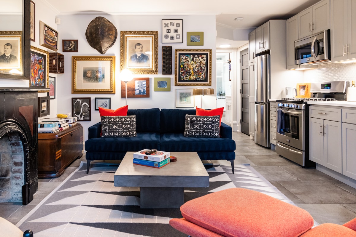 The living area features a plush blue sofa adorned with decorative cushions, set against a gallery wall of framed art. A contemporary coffee table sits centrally, with a modern armchair nearby. The kitchen area showcases sleek cabinetry and stainless steel appliances, enhancing the cozy ambiance.