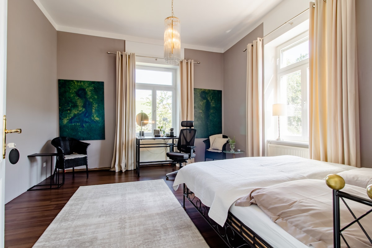 A spacious bedroom features a large bed with crisp linens and two vibrant green artwork pieces on the walls. Natural light floods the room through two large windows, complemented by soft curtains. A chair and a desk with a mirror complete the room's functional layout.