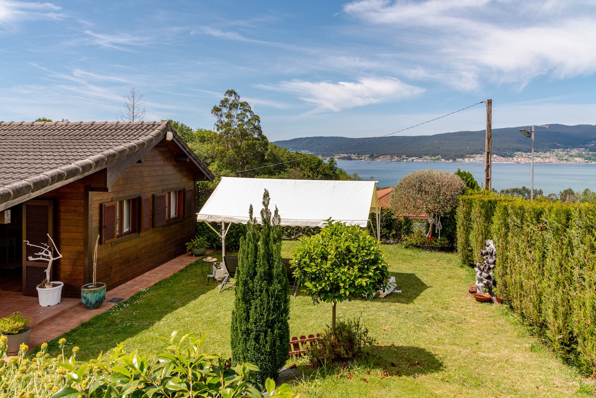 A wooden cabin is nestled within a spacious garden, featuring lush greenery and flowering plants. A tent is set up in the yard, providing outdoor dining space. The view encompasses a serene body of water, framed by trees under a clear blue sky.