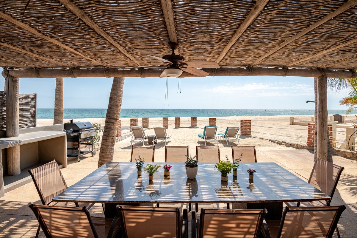 An outdoor dining area features a large table surrounded by chairs, shaded by a thatched roof. A barbecue grill is positioned to one side, while the sandy beach and ocean are visible in the background, creating a serene coastal setting.