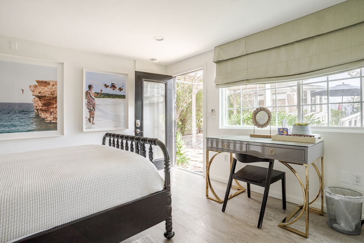 A bright bedroom features a spacious bed with a neutral coverlet, complemented by a large window and a door leading to the outdoor area. Natural light floods the room, highlighting a sleek desk with a decorative mirror and a view of lush greenery outside.
